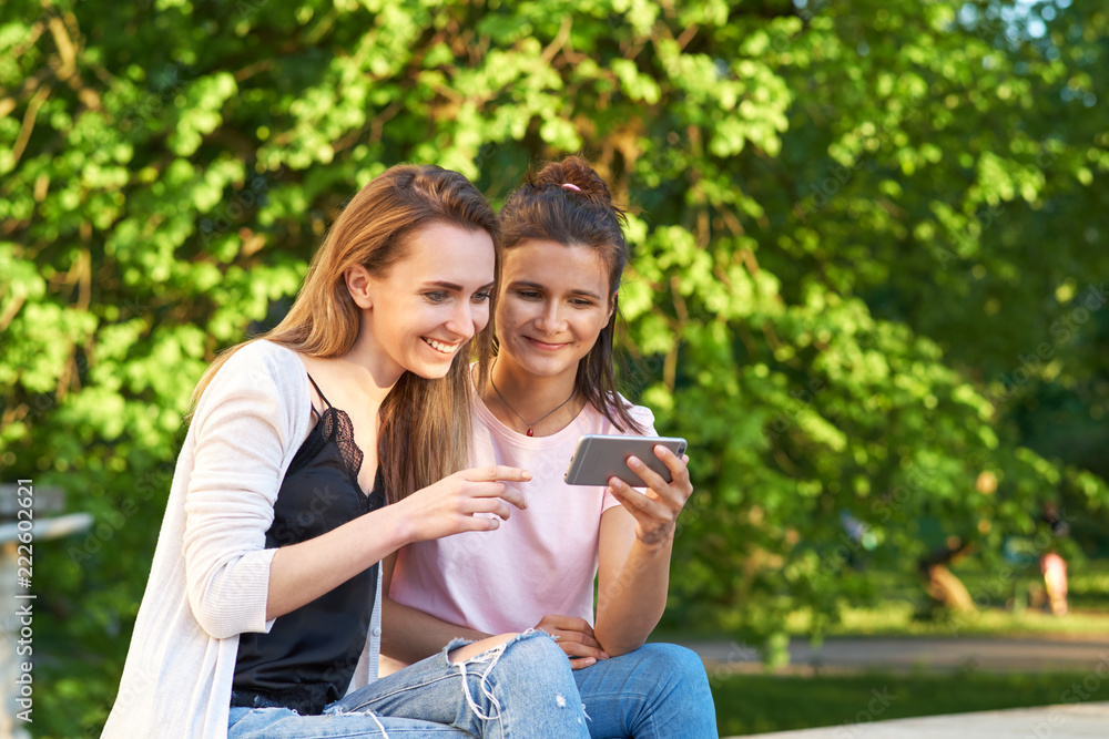 Two young girls are sitting in the Park on the background of trees and watching photos on smartphone. The concept of mobile devices, gadgets, wireless communication. Lifestyle and outdoor recreation Two young girls are sitting in the Park on the background of trees and watching photos on smartphone. The concept of mobile devices, gadgets, wireless communication. Lifestyle and outdoor recreation