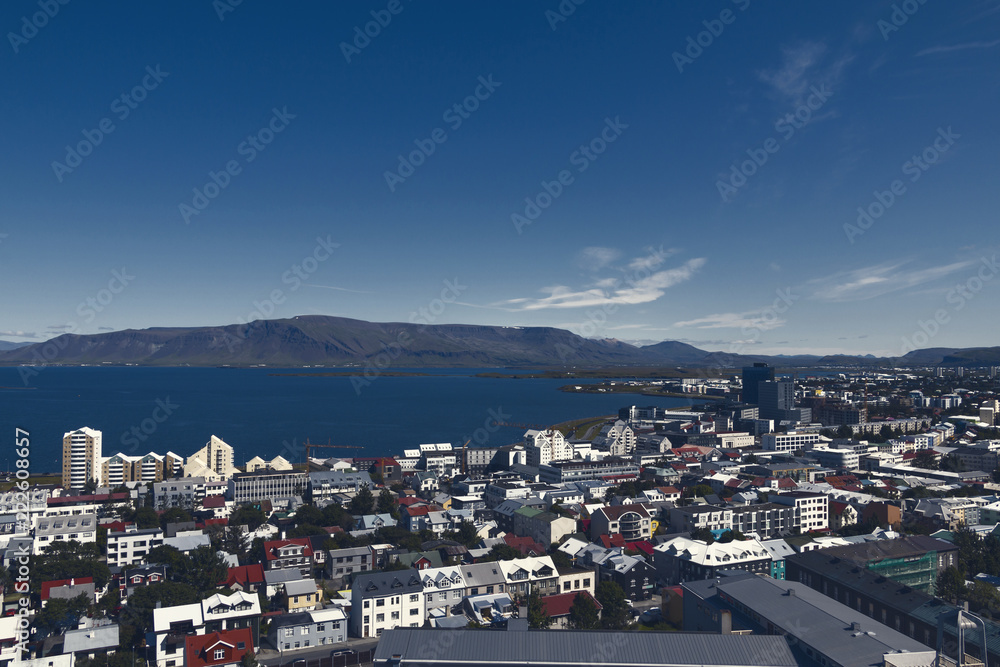 Aerial view on capital of Iceland - city of Reykjavik - ocean bay, hills, mountains, green medows, roadways, hills, streets, houses, traffic, roofing - on sunny day.