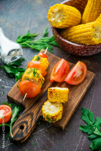 Grilled corn on skewers with tomatoes, herbs and spices on a dark background.
