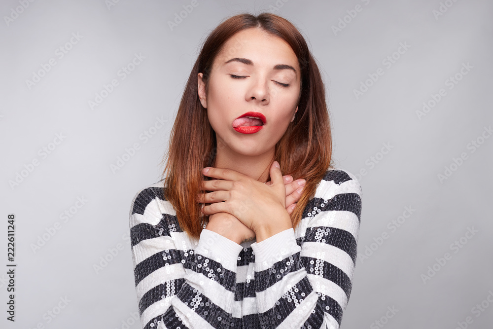 Attractive female in despair makes suicide gestures and smothers herself, suffers from suffocation, wears striped t shirt, has tongue out, poses against gray background. People, body language concept. Attractive female in despair makes suicide gestures and smothers herself, suffers from suffocation, wears striped t shirt, has tongue out, poses against gray background. People, body language concept.
