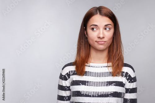 Close-up portrait of young female with pure skin, wearing glitter sweater with stripes, looking suspicious aside, posing against gray studio wall with copy space for advertising. People, lifestyle.
