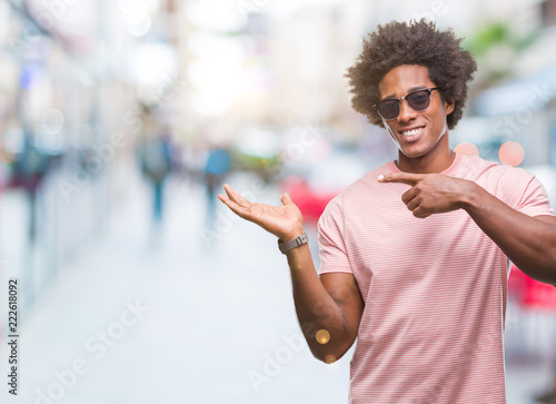 Afro american man wearing sunglasses over isolated background amazed and smiling to the camera while presenting with hand and pointing with finger.