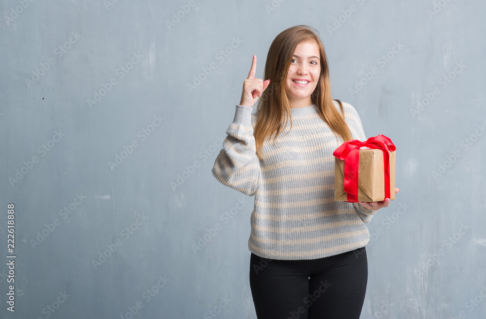 Young adult woman over grey grunge wall holding a present surprised with an idea or question pointing finger with happy face, number one Young adult woman over grey grunge wall holding a present surprised with an idea or question pointing finger with happy face, number one