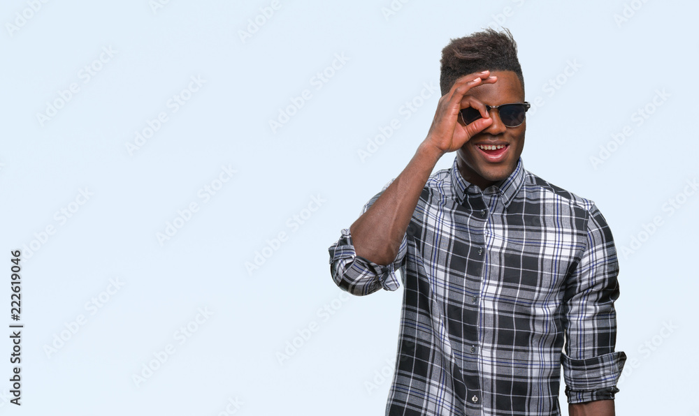 Naklejka premium Young african american man wearing sunglasses over isolated background doing ok gesture with hand smiling, eye looking through fingers with happy face.