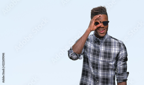 Young african american man wearing sunglasses over isolated background doing ok gesture with hand smiling, eye looking through fingers with happy face.