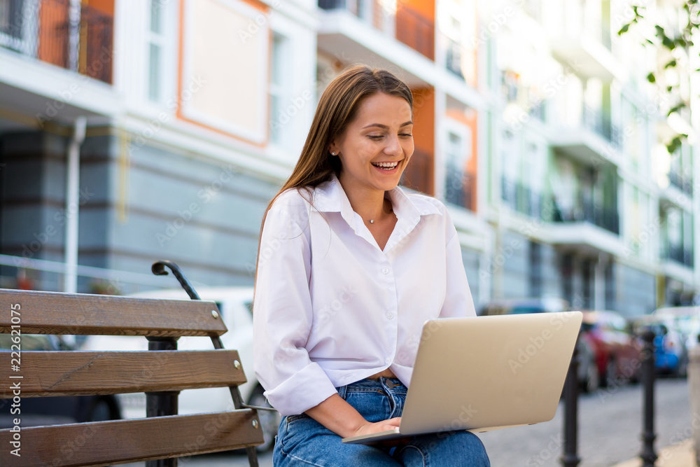 Young beautiful laughing female with a laptop sits on a bench in the business part of the city. Young beautiful laughing female with a laptop sits on a bench in the business part of the city.