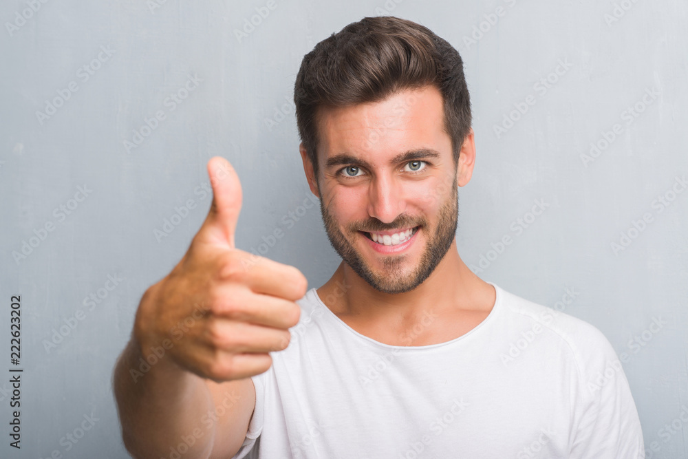Handsome young man over grey grunge wall happy with big smile doing ok sign, thumb up with fingers, excellent sign Handsome young man over grey grunge wall happy with big smile doing ok sign, thumb up with fingers, excellent sign
