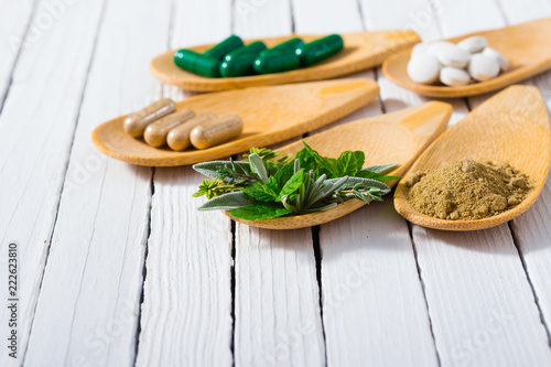 herbal leaves, ground herb powder and medicament pills on bamboo spoons, white wooden table