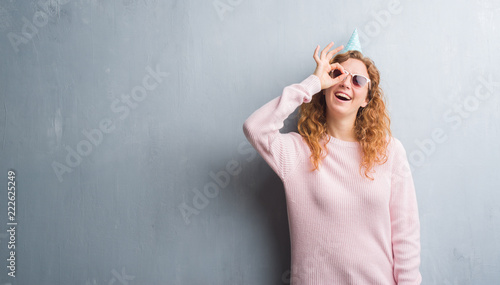 Young redhead woman over grey grunge wall wearing birthday cap with happy face smiling doing ok sign with hand on eye looking through fingers