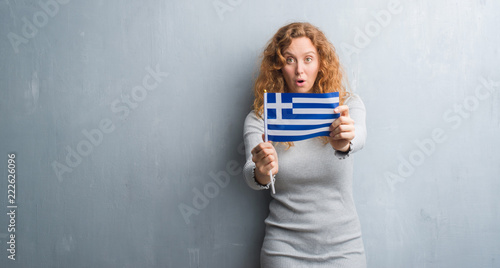 Young redhead woman over grey grunge wall holding flag of Greece scared in shock with a surprise face, afraid and excited with fear expression