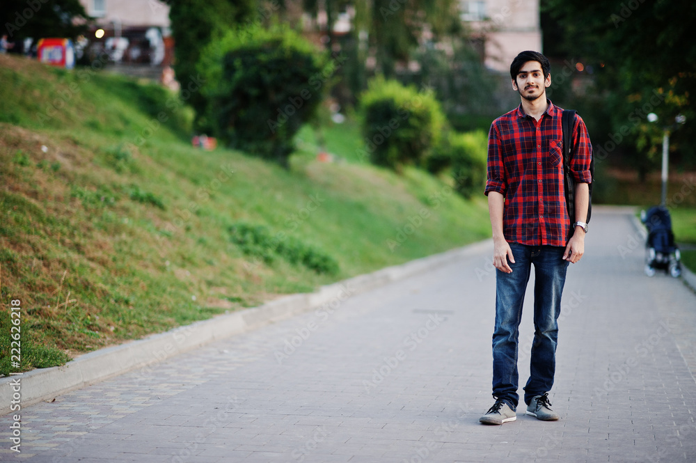 Young indian student man at red checkered shirt and jeans with backpack posed at street. Young indian student man at red checkered shirt and jeans with backpack posed at street.