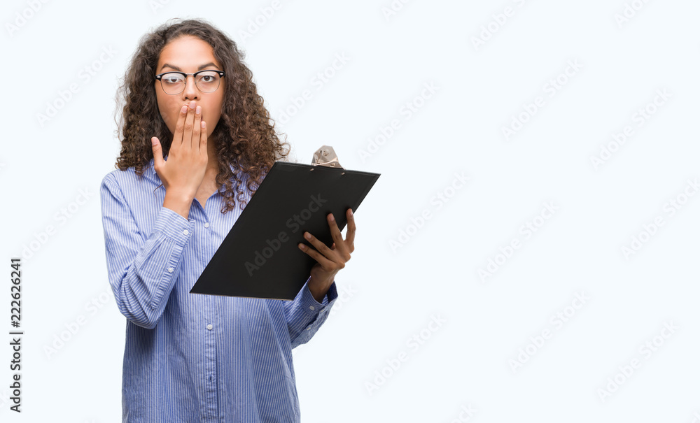 Fototapeta premium Young hispanic business woman holding clipboard cover mouth with hand shocked with shame for mistake, expression of fear, scared in silence, secret concept