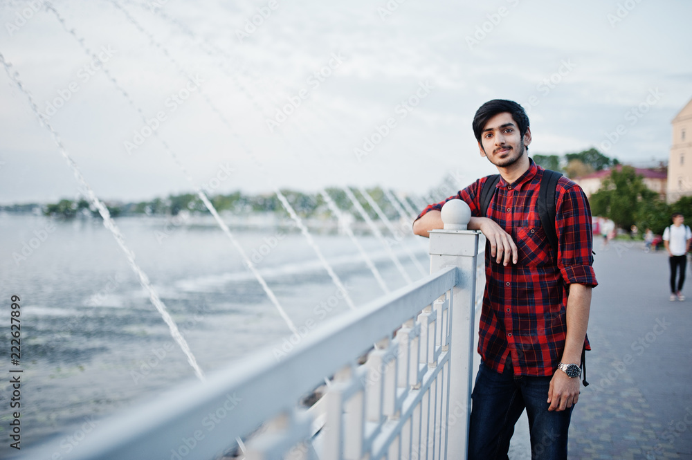 Young indian student man at checkered shirt and jeans with backpack posed on evening city against fountains. Young indian student man at checkered shirt and jeans with backpack posed on evening city against fountains.