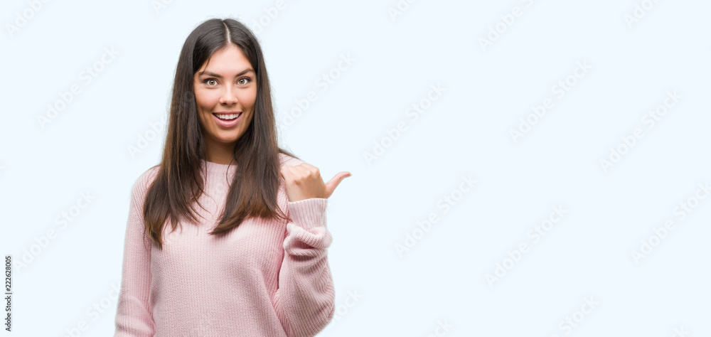 Young beautiful hispanic woman wearing a sweater smiling with happy face looking and pointing to the side with thumb up. Young beautiful hispanic woman wearing a sweater smiling with happy face looking and pointing to the side with thumb up.