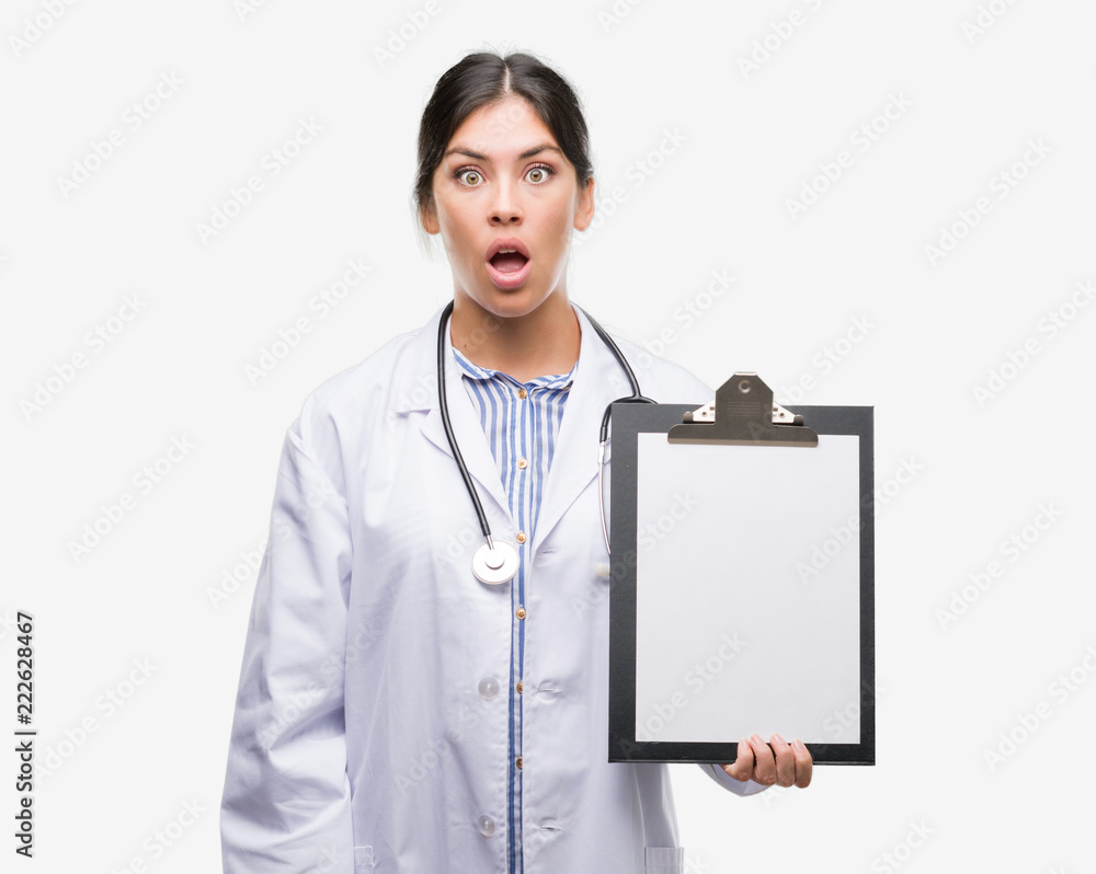 Young hispanic doctor woman holding clipboard scared in shock with a surprise face, afraid and excited with fear expression Young hispanic doctor woman holding clipboard scared in shock with a surprise face, afraid and excited with fear expression