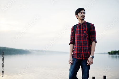 Young indian student man at checkered shirt and jeans with backpack posed on evening city against lake. © AS Photo Family