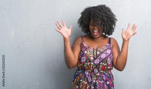 Obraz na plátně Young african american woman over grey grunge wall wearing colorful dress celebrating mad and crazy for success with arms raised and closed eyes screaming excited