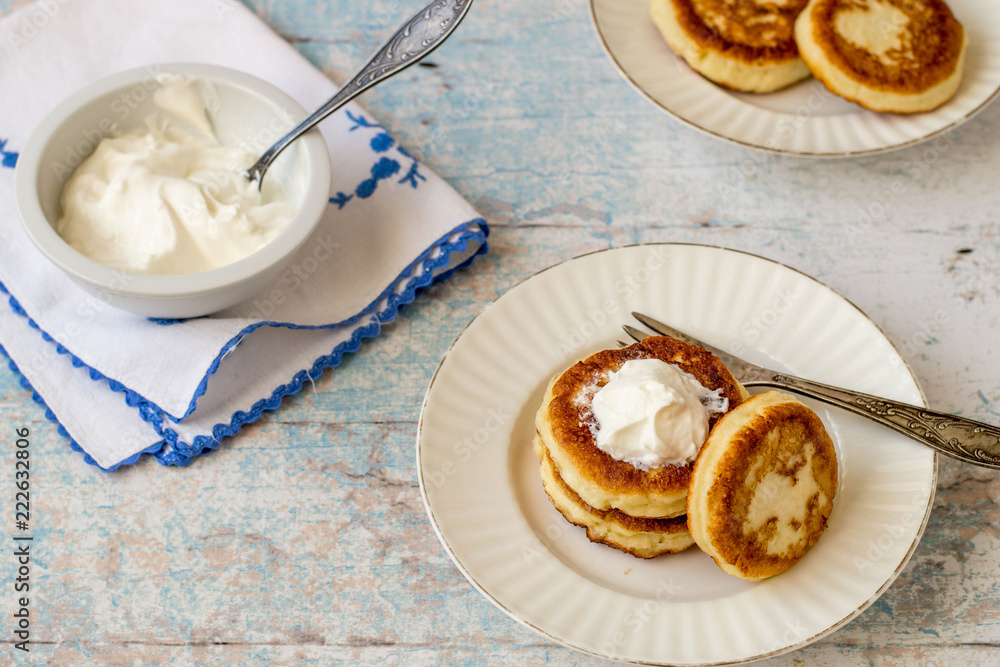 Delicious fried syrniki on a round plate, next to a sour cream cup on a cotton napkin on an old wooden table. Delicious fried syrniki on a round plate, next to a sour cream cup on a cotton napkin on an old wooden table.