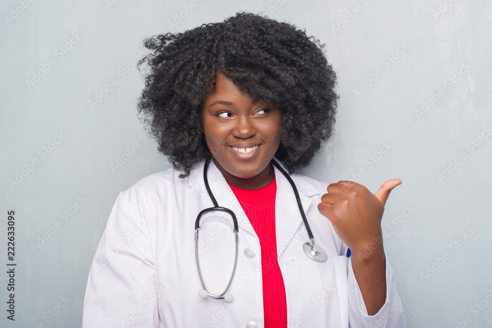 Young african american doctor woman over grey grunge wall smiling with happy face looking and pointing to the side with thumb up. Young african american doctor woman over grey grunge wall smiling with happy face looking and pointing to the side with thumb up.