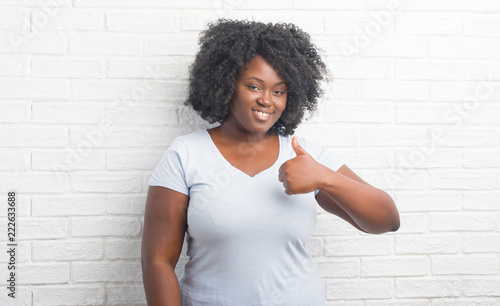 Young african american plus size woman over white brick wall doing happy thumbs up gesture with hand. Approving expression looking at the camera with showing success.