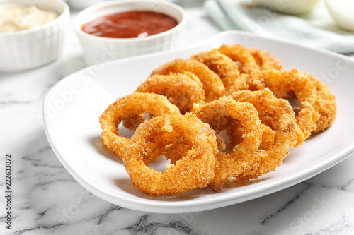 Plate with homemade crunchy fried onion rings and sauce on marble table, closeup