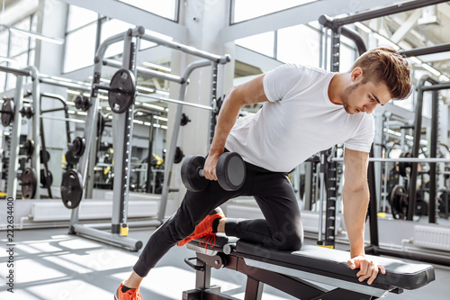 Handsome caucasian man doing biceps lifting with dumbbell on bench in crossfit studio