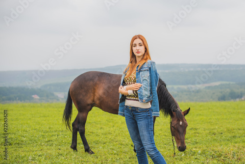 Portrait of young beautiful woman posing at meadow, near horse. Model wearing stylish blue denim jacket and jeans, warm sweater. Female fashion concept. 