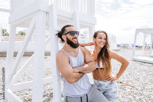 Two close best friends wearing sportive clothes, listening to the music with smartphone while walking together at seaside in summer morning, feeling more than friendship. photo