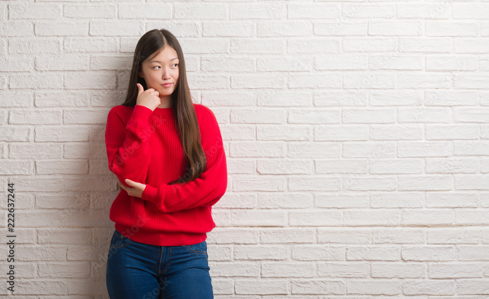 Young Chinese woman over brick wall with hand on chin thinking about question, pensive expression. Smiling with thoughtful face. Doubt concept. Young Chinese woman over brick wall with hand on chin thinking about question, pensive expression. Smiling with thoughtful face. Doubt concept.