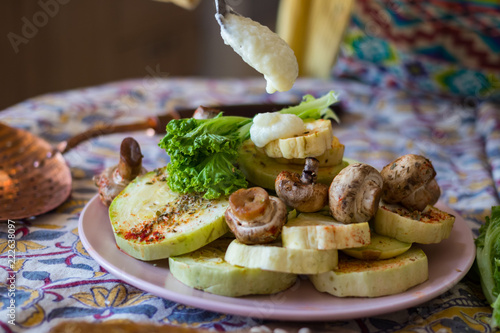 Woman hands adds toum garlic sauce paste, vegan mayonnaise with tea spoon to  backed, roasted assorted vegetables on plate. Healthy vegan lunch or vegetarian dinner. 