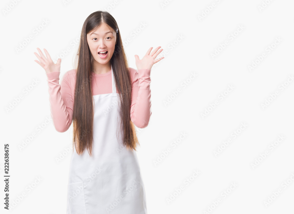 Young Chinese shop owner woman over isolated background wearing white apron very happy and excited, winner expression celebrating victory screaming with big smile and raised hands Young Chinese shop owner woman over isolated background wearing white apron very happy and excited, winner expression celebrating victory screaming with big smile and raised hands