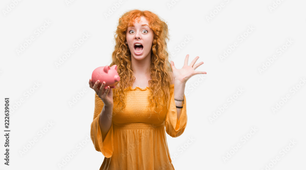 Young redhead woman holding piggy bank very happy and excited, winner expression celebrating victory screaming with big smile and raised hands Young redhead woman holding piggy bank very happy and excited, winner expression celebrating victory screaming with big smile and raised hands