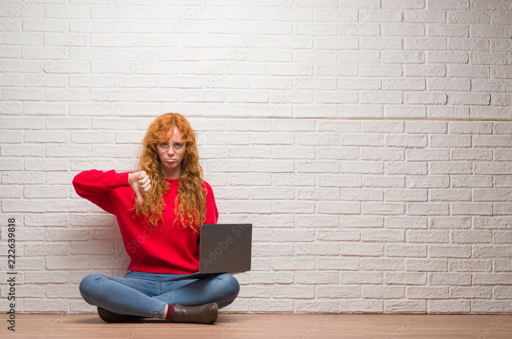 Young redhead woman sitting over brick wall using computer laptop with angry face, negative sign showing dislike with thumbs down, rejection concept Young redhead woman sitting over brick wall using computer laptop with angry face, negative sign showing dislike with thumbs down, rejection concept