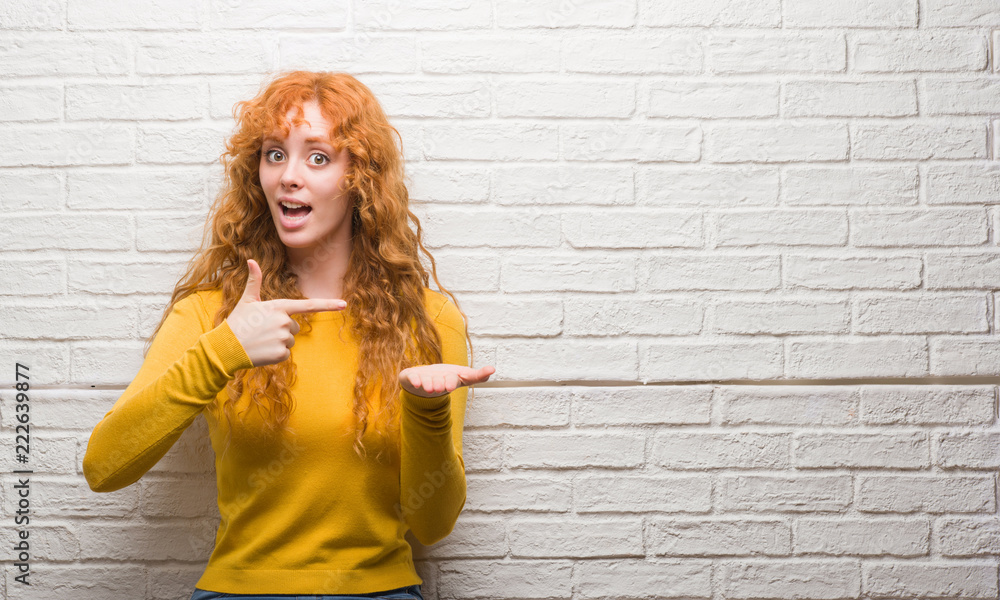 Young redhead woman standing over brick wall very happy pointing with hand and finger Young redhead woman standing over brick wall very happy pointing with hand and finger