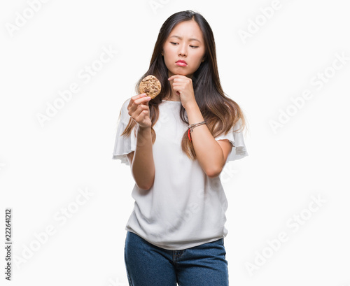 Young asian woman eating chocolate chip cookie over isolated background serious face thinking about question, very confused idea