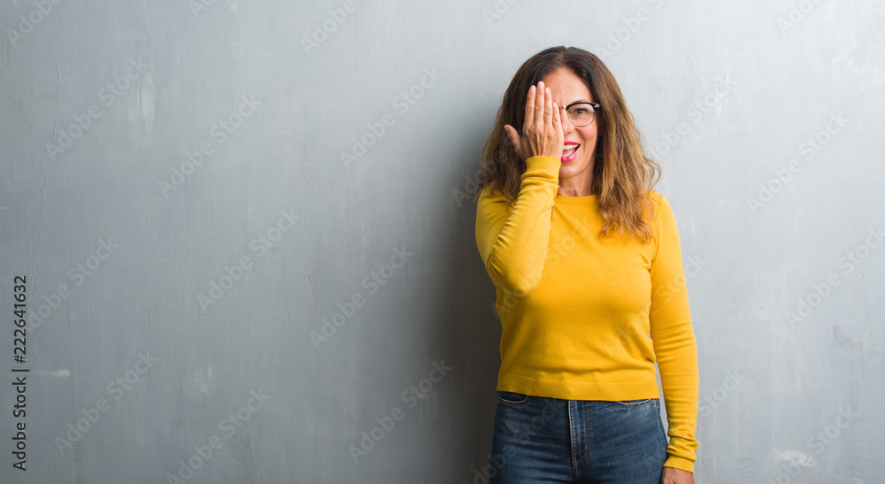 Middle age hispanic woman over grey wall wearing glasses covering one eye with hand with confident smile on face and surprise emotion.