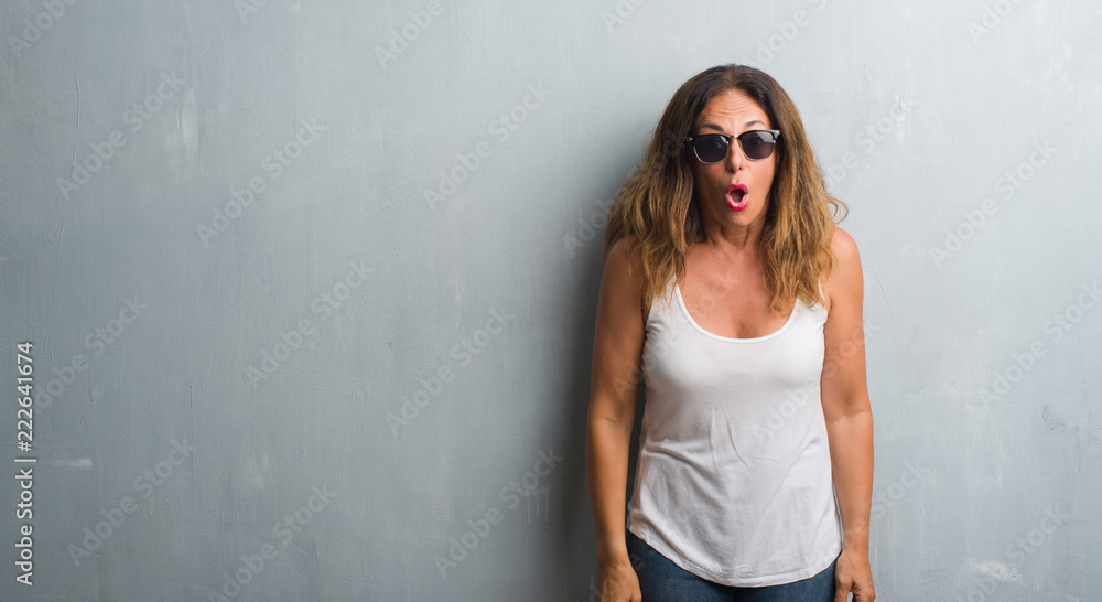 Middle age hispanic woman over grey wall wearing sunglasses afraid and shocked with surprise expression, fear and excited face. Middle age hispanic woman over grey wall wearing sunglasses afraid and shocked with surprise expression, fear and excited face.