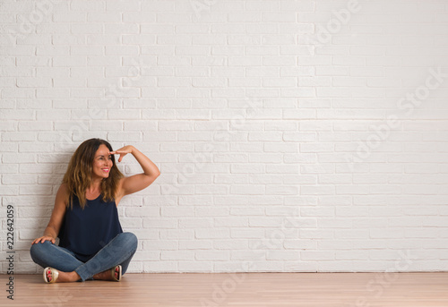 Middle age hispanic woman sitting on the floor over white brick wall very happy and smiling looking far away with hand over head. Searching concept.