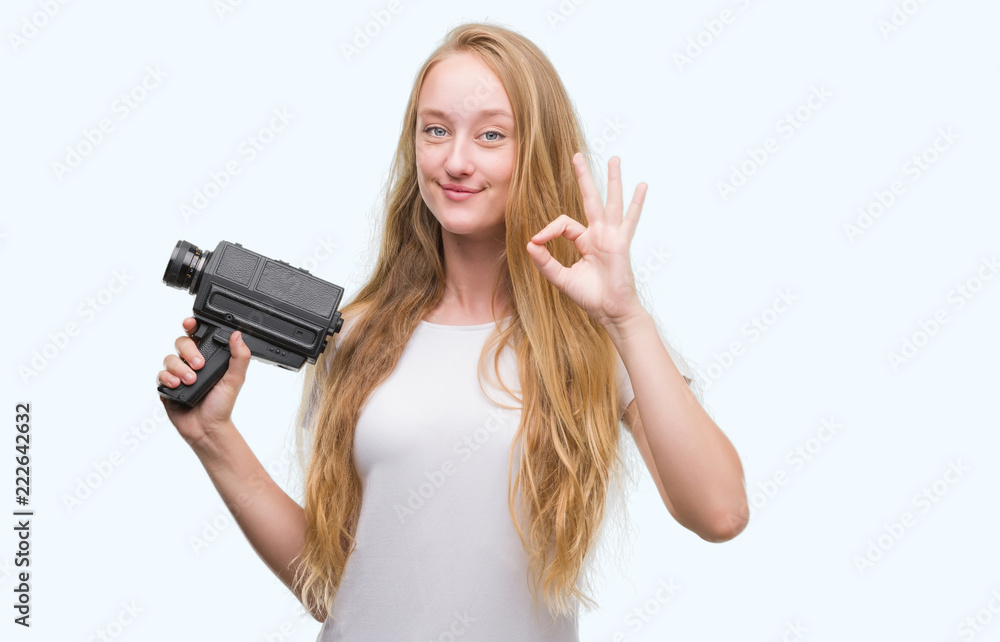 Blonde woman teenager filming holding super 8 video camera doing ok sign with fingers, excellent symbol Blonde woman teenager filming holding super 8 video camera doing ok sign with fingers, excellent symbol