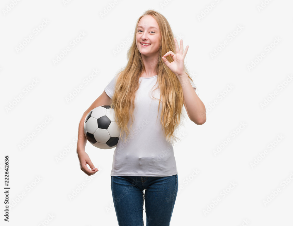 Blonde teenager woman holding soccer football ball doing ok sign with fingers, excellent symbol Blonde teenager woman holding soccer football ball doing ok sign with fingers, excellent symbol