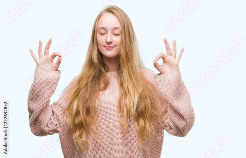 Blonde teenager woman wearing pink sweater relax and smiling with eyes closed doing meditation gesture with fingers. Yoga concept.