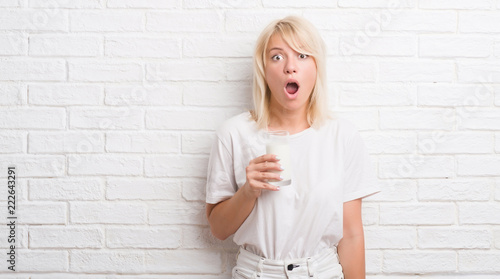Adult caucasian woman over white brick wall drinking glass of milk scared in shock with a surprise face, afraid and excited with fear expression