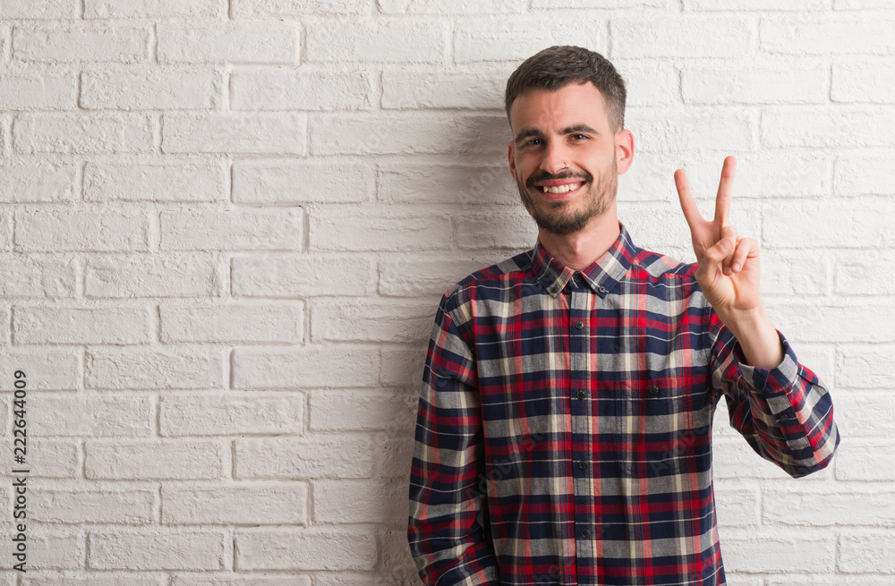 Young adult man standing over white brick wall smiling looking to the camera showing fingers doing victory sign. Number two. Young adult man standing over white brick wall smiling looking to the camera showing fingers doing victory sign. Number two.