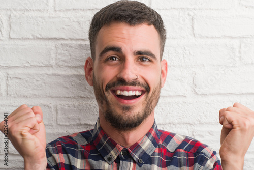 Young adult man standing over white brick wall screaming proud and celebrating victory and success very excited, cheering emotion