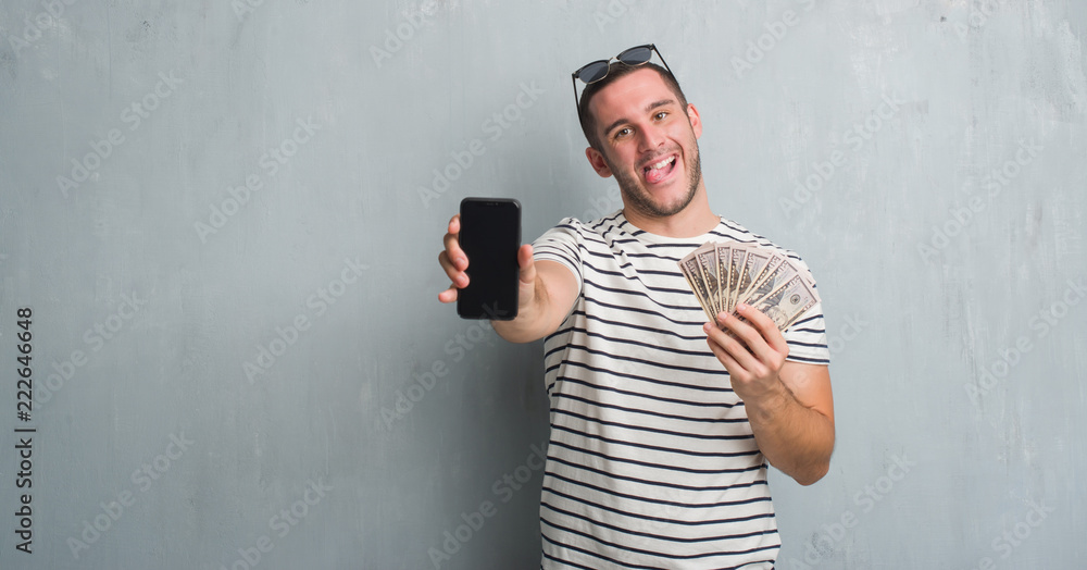 Fototapeta premium Young caucasian man over grey grunge wall holding dollars and smarphone sticking tongue out happy with funny expression. Emotion concept.