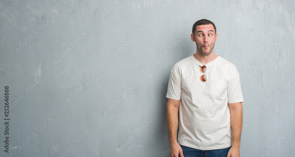 Young caucasian man over grey grunge wall wearing sunglasses making fish face with lips, crazy and comical gesture. Funny expression. Young caucasian man over grey grunge wall wearing sunglasses making fish face with lips, crazy and comical gesture. Funny expression.