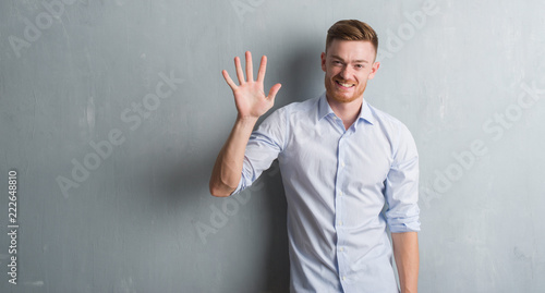 Young redhead business man over grey grunge wall showing and pointing up with fingers number five while smiling confident and happy. © Krakenimages.com