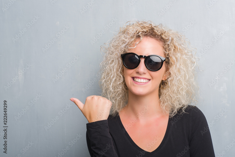 Young blonde woman with curly hair over grunge grey background smiling with happy face looking and pointing to the side with thumb up. Young blonde woman with curly hair over grunge grey background smiling with happy face looking and pointing to the side with thumb up.