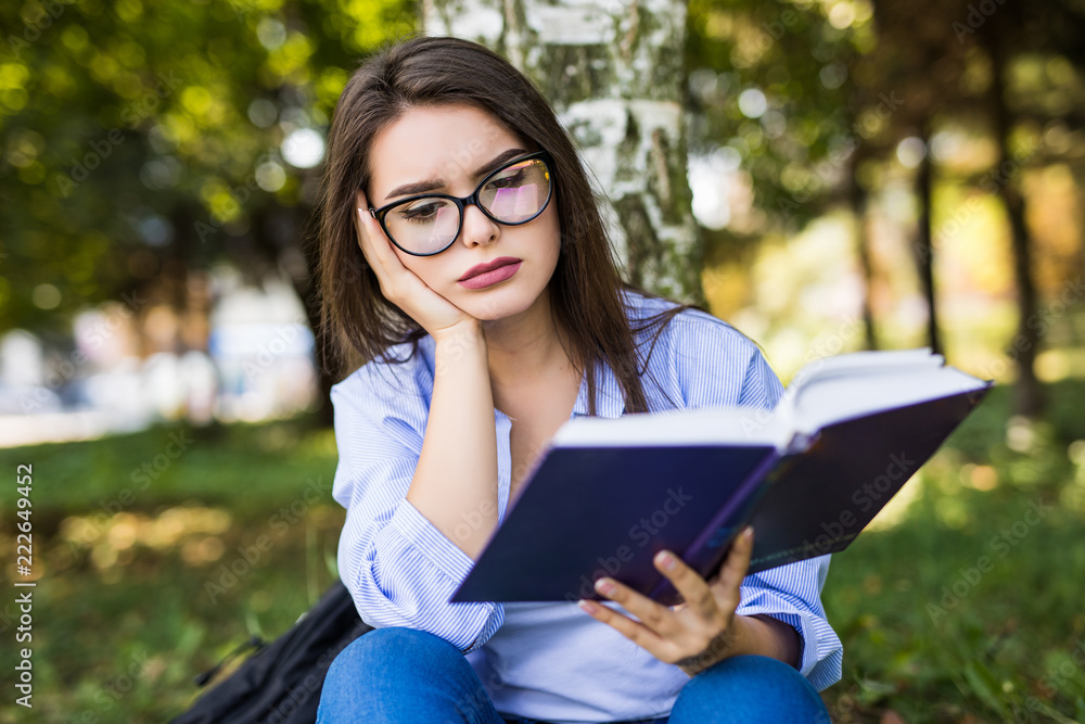 Young woman in eyeglasses sitting on grass under tree and reading book in park Young woman in eyeglasses sitting on grass under tree and reading book in park