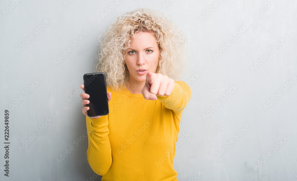 Young blonde woman over grunge grey background showing blank screen of smartphone pointing with finger to the camera and to you, hand sign, positive and confident gesture from the front Young blonde woman over grunge grey background showing blank screen of smartphone pointing with finger to the camera and to you, hand sign, positive and confident gesture from the front